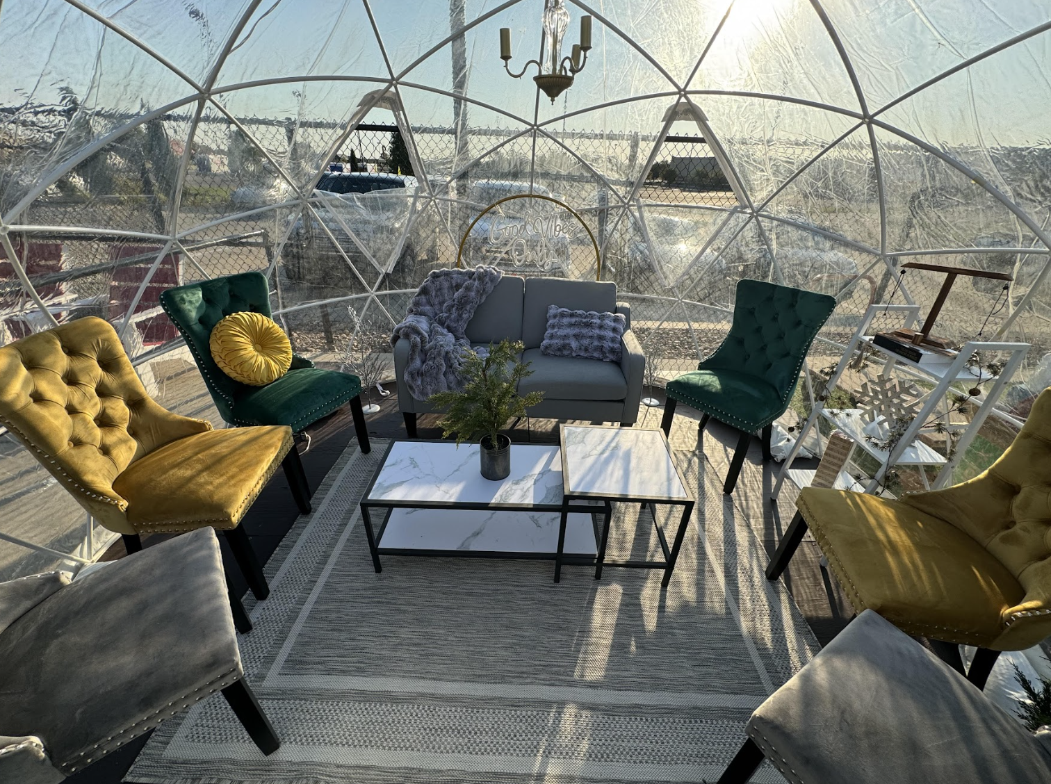 A cozy seating area inside a clear geodesic dome features a gray sofa with pillows, green and yellow tufted chairs, a white coffee table with a plant, and sunlight streaming through the transparent walls.